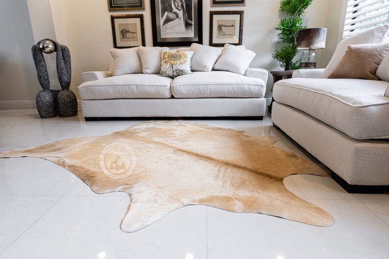 May include: A light brown cowhide rug with a natural, irregular shape. The rug is laid out on a white tiled floor in front of a white sofa and a white armchair.