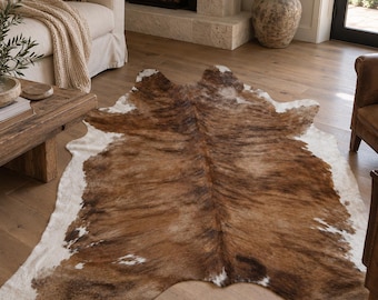 Pode incluir: Um tapete de pele de vaca com um padrão natural de pelo castanho, bege e branco. O tapete tem uma forma orgânica irregular com uma faixa central castanha escura e manchas brancas mais claras ao longo das bordas sobre um piso de madeira.