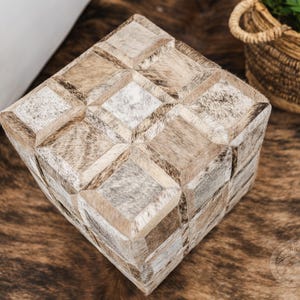 K&ouml;nnte beinhalten: Ein quadratischer Hocker mit einem geometrischen Muster aus verschiedenen Braunt&ouml;nen und wei&szlig;em Leder. Der Hocker steht auf einem braunen und wei&szlig;en Kuhfellteppich.