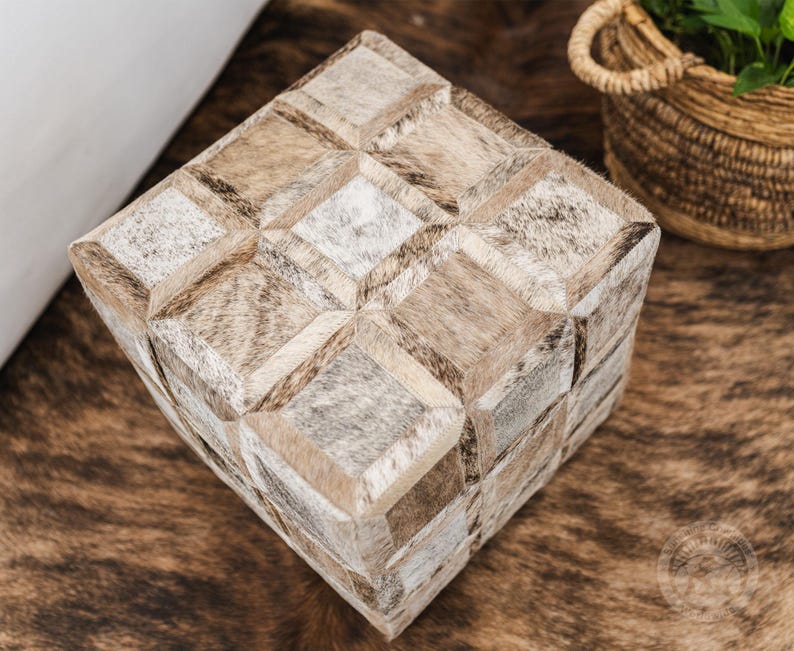 K&ouml;nnte beinhalten: Ein quadratischer Hocker mit einem geometrischen Muster aus verschiedenen Braunt&ouml;nen und wei&szlig;em Leder. Der Hocker steht auf einem braunen und wei&szlig;en Kuhfellteppich.
