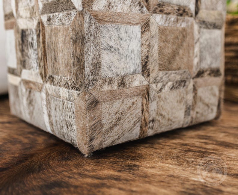 K&ouml;nnte beinhalten: Ein quadratischer Hocker mit einem geometrischen Muster aus verschiedenen Braunt&ouml;nen und wei&szlig;em Kuhfell. Der Hocker steht auf einem braunen Kuhfellteppich.