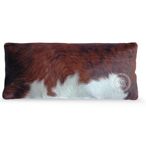 May include: A rectangular throw pillow covered in genuine cowhide featuring a natural tri-color pattern of brown, black, and white patches. The surface has a textured, hair-on-hide finish with a circular logo stamp visible on the lower right side.