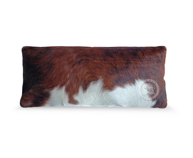 May include: A rectangular throw pillow covered in genuine cowhide featuring a natural tri-color pattern of brown, black, and white patches. The surface has a textured, hair-on-hide finish with a circular logo stamp visible on the lower right side.