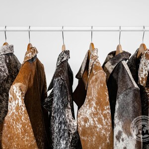 May include: A collection of cowhide jackets in various colors and patterns hanging on wooden hangers. The jackets feature brown, black, and white spotted designs. The jackets are displayed on a white metal rod against a white background.