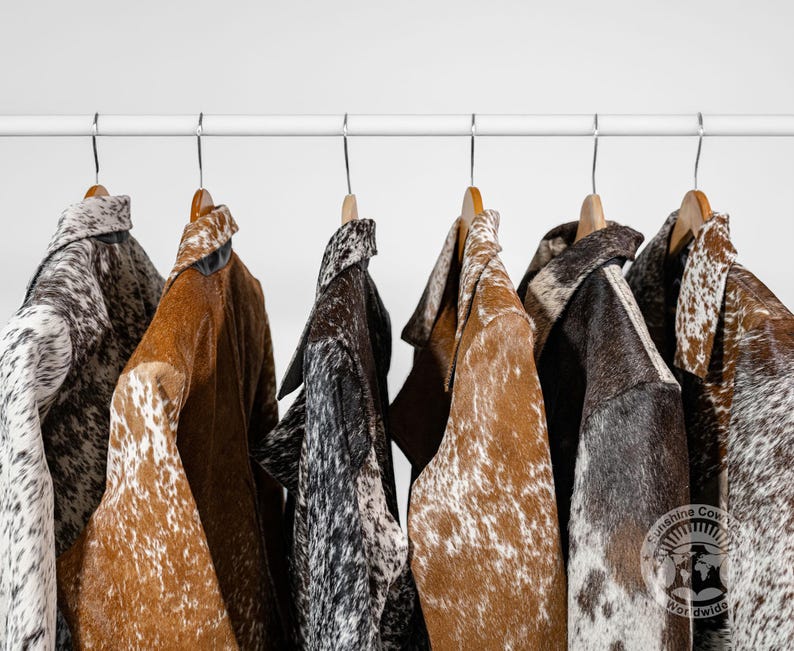 May include: A collection of cowhide jackets in various colors and patterns hanging on wooden hangers. The jackets feature brown, black, and white spotted designs. The jackets are displayed on a white metal rod against a white background.