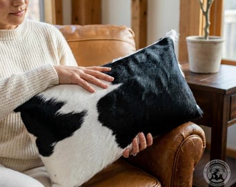 May include: A cowhide pillow with a black and white pattern. The pillow is rectangular and appears to be soft. The pillow is being held by a person wearing a cream-colored sweater. The pillow is on a brown leather chair.