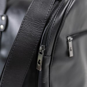May include: A black leather duffel bag featuring a smooth texture, a thick shoulder strap attached with silver-toned rivets, and a side zippered pocket. The metal zipper pulls are rectangular with a small cutout detail at the top of each pull.