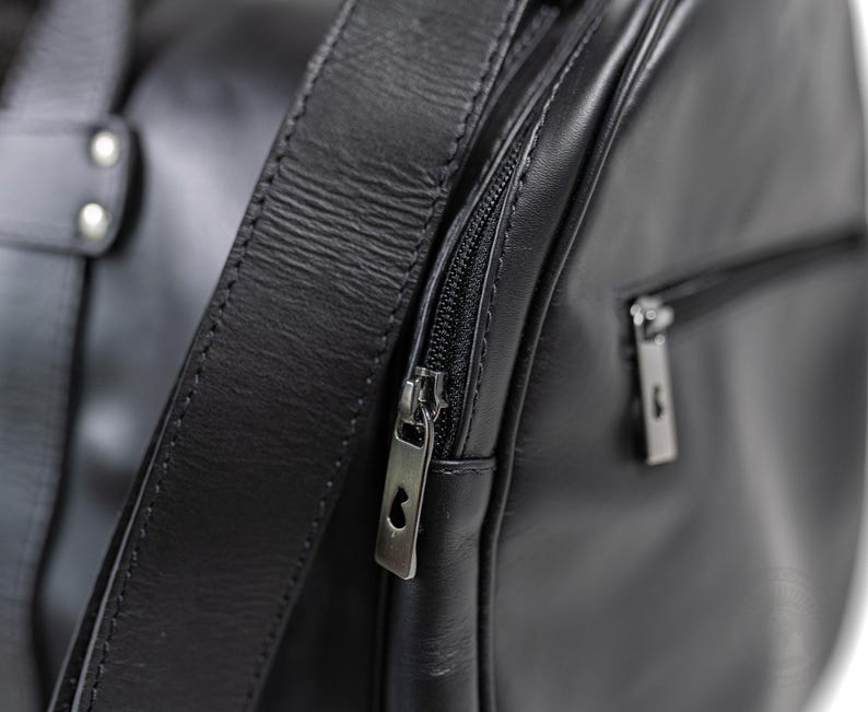 May include: A black leather duffel bag featuring a smooth texture, a thick shoulder strap attached with silver-toned rivets, and a side zippered pocket. The metal zipper pulls are rectangular with a small cutout detail at the top of each pull.