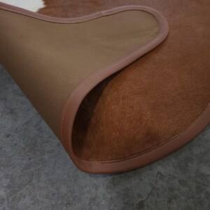 May include: A brown and white cowhide rug with a leather border. The rug's underside is a tan fabric. The rug is partially rolled up, revealing the underside and the texture of the cowhide.