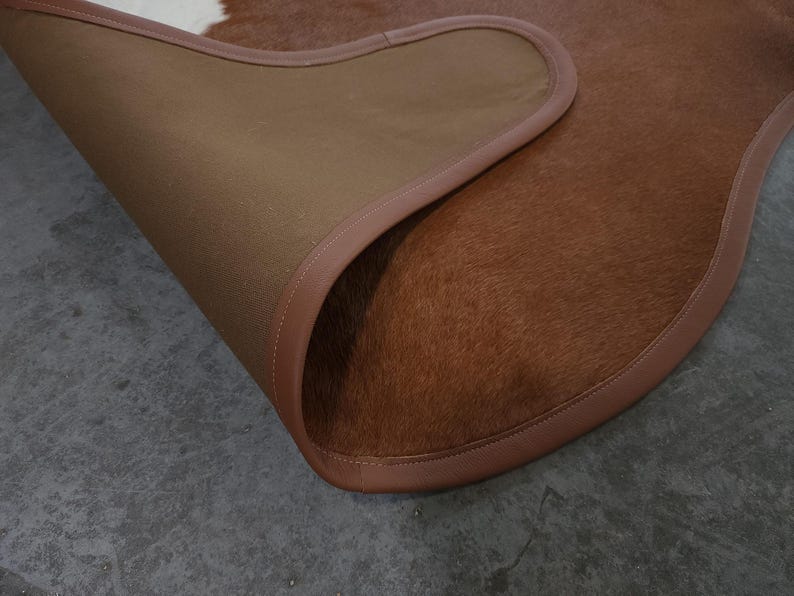 May include: A brown and white cowhide rug with a leather border. The rug's underside is a tan fabric. The rug is partially rolled up, revealing the underside and the texture of the cowhide.