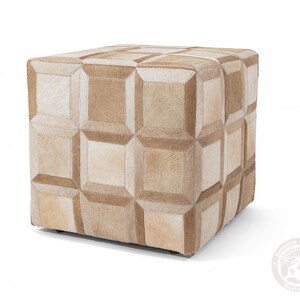 May include: A square ottoman with a patchwork design. The ottoman is covered in sections of tan and cream-colored cowhide, stitched together to create a geometric pattern. The overall effect is a neutral-toned, textured piece of furniture.