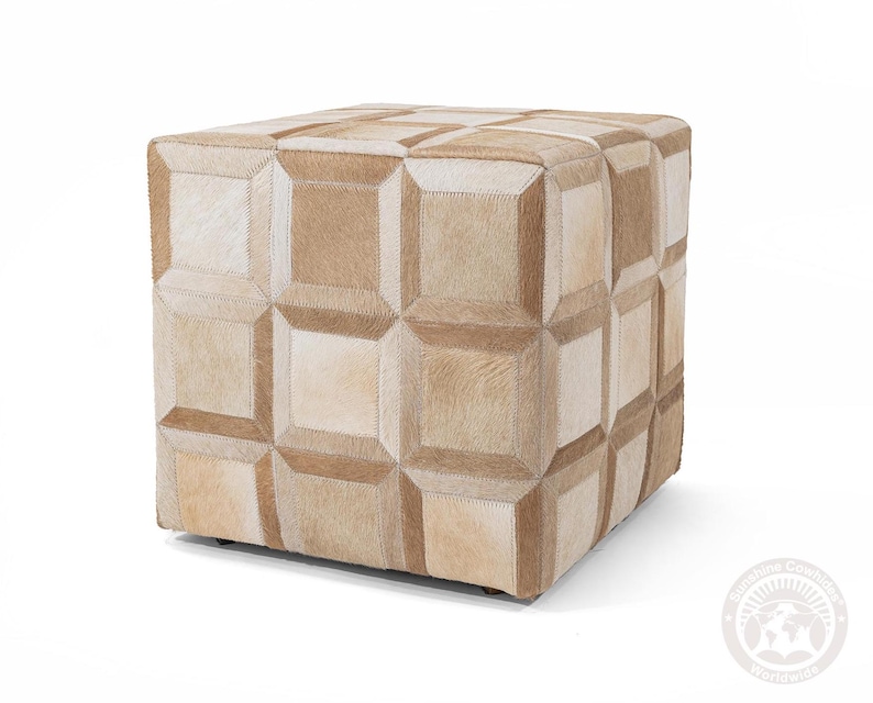 May include: A square ottoman with a patchwork design. The ottoman is covered in sections of tan and cream-colored cowhide, stitched together to create a geometric pattern. The overall effect is a neutral-toned, textured piece of furniture.