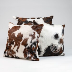 May include: Three square throw pillows featuring a cowhide pattern with patches of brown, white, and black fur. The pillows have a textured, hair-on-hide appearance. One pillow displays a circular logo with the text Sun Hide Country Worldwide.