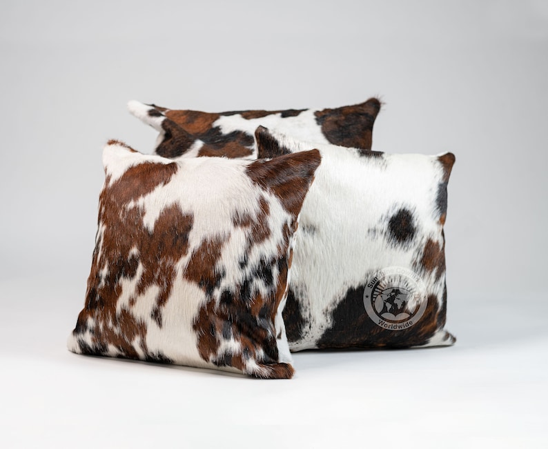 May include: Three square throw pillows featuring a cowhide pattern with patches of brown, white, and black fur. The pillows have a textured, hair-on-hide appearance. One pillow displays a circular logo with the text Sun Hide Country Worldwide.