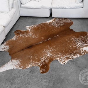 May include: A brown and white cowhide rug with an irregular shape. The rug has a natural pattern with a mix of brown and white spots. The rug is placed on a grey concrete floor, with white sofas in the background.