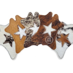 May include: Six cowhide coasters in a range of colours, including white, brown, and tan. Each coaster has a stitched white star and a stitched border. The coasters are arranged in a fan shape.