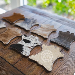May include: A selection of cowhide coasters in shades of brown, grey, and black and white. The coasters are displayed on a wooden table, highlighting their individual patterns and textures. One coaster features a logo.