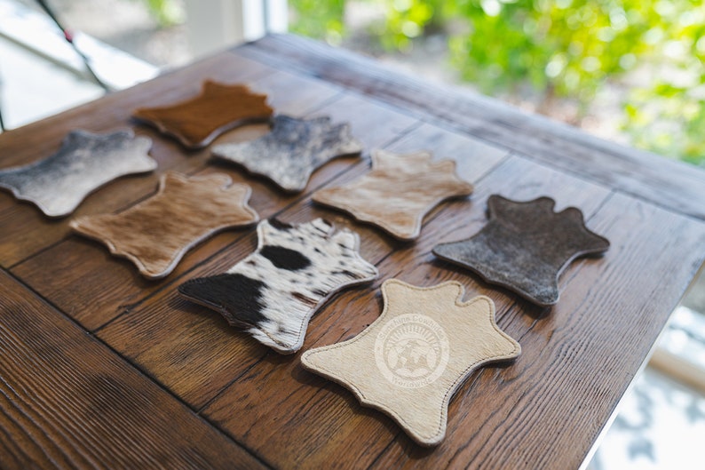 May include: A selection of cowhide coasters in shades of brown, grey, and black and white. The coasters are displayed on a wooden table, highlighting their individual patterns and textures. One coaster features a logo.