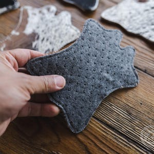 May include: A hand holding a grey, star-shaped felt coaster with small black dots. Other coasters with cowhide patterns are visible in the background, resting on a wooden surface. The felt coaster is designed to protect surfaces from heat and moisture.