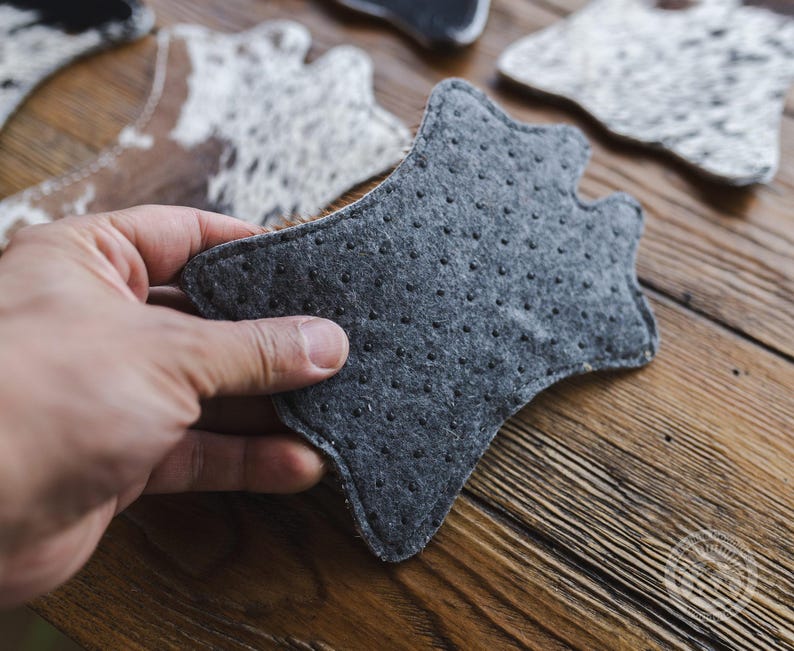 May include: A hand holding a grey, star-shaped felt coaster with small black dots. Other coasters with cowhide patterns are visible in the background, resting on a wooden surface. The felt coaster is designed to protect surfaces from heat and moisture.