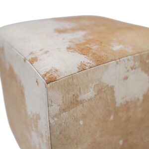 May include: A square ottoman upholstered in genuine cowhide leather featuring a natural mottled pattern of white and light tan patches. The cube-shaped furniture piece has clean, straight edges and a soft, textured surface with visible hair.