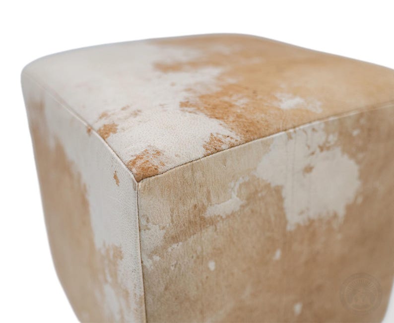 May include: A square ottoman upholstered in genuine cowhide leather featuring a natural mottled pattern of white and light tan patches. The cube-shaped furniture piece has clean, straight edges and a soft, textured surface with visible hair.