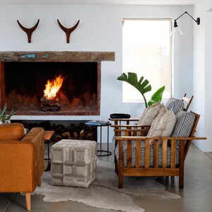 May include: A cozy living room scene with a fireplace, two wooden armchairs, and a leather sofa. The fireplace has a burning fire inside. A patterned ottoman and a cowhide rug add texture to the space. Decorative antlers hang above the fireplace.