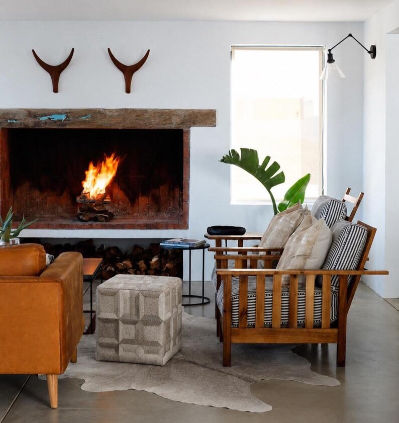 May include: A cozy living room scene with a fireplace, two wooden armchairs, and a leather sofa. The fireplace has a burning fire inside. A patterned ottoman and a cowhide rug add texture to the space. Decorative antlers hang above the fireplace.
