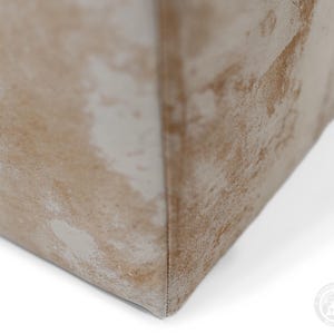 May include: A close-up view of a corner of a rectangular object featuring a textured surface with a mottled pattern in shades of beige, tan, and off-white. The material appears soft and fabric-like, with a visible vertical seam running along the edge.