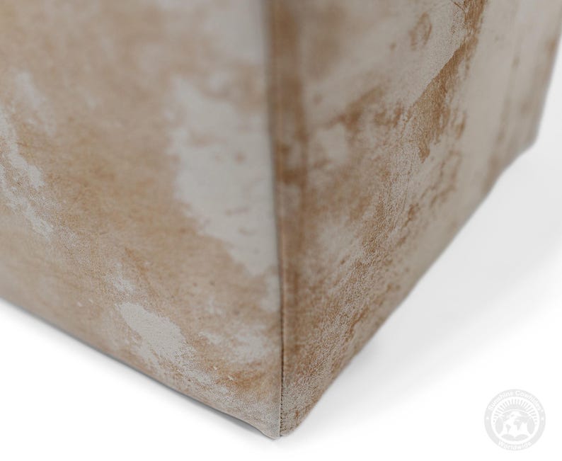 May include: A close-up view of a corner of a rectangular object featuring a textured surface with a mottled pattern in shades of beige, tan, and off-white. The material appears soft and fabric-like, with a visible vertical seam running along the edge.