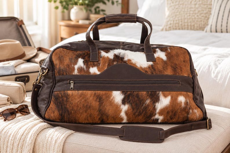 May include: A duffel bag crafted from dark brown leather and brown, white, and black cowhide fur. The bag features two top handles, a detachable shoulder strap with a padded section, and a horizontal zippered pocket across the front panel.