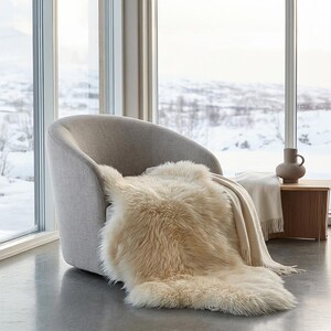 Puede incluir: Una escena interior acogedora presenta un sill&oacute;n gris claro con una manta de piel sint&eacute;tica color crema. Una manta beige con flecos y una peque&ntilde;a mesa auxiliar de madera con un jarr&oacute;n completan la escena. El fondo muestra un paisaje nevado.
