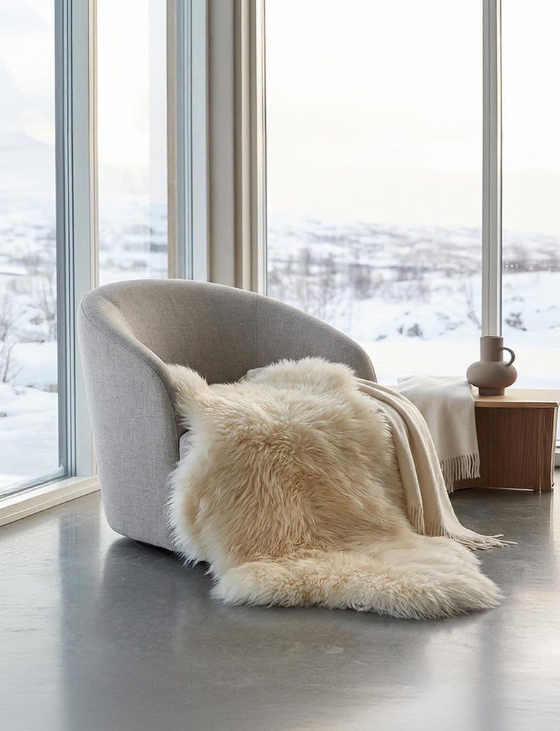 Puede incluir: Una escena interior acogedora presenta un sill&oacute;n gris claro con una manta de piel sint&eacute;tica color crema. Una manta beige con flecos y una peque&ntilde;a mesa auxiliar de madera con un jarr&oacute;n completan la escena. El fondo muestra un paisaje nevado.