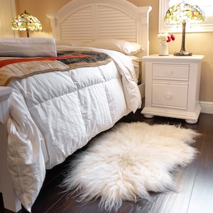 Puede incluir: Un dormitorio con una cama blanca, una manta estampada y una alfombra blanca y esponjosa sobre un suelo de madera oscura. Una mesita de noche blanca con una l&aacute;mpara y una pantalla de vidrieras est&aacute; junto a la cama. Ambiente acogedor.