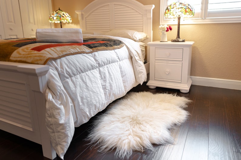 Puede incluir: Un dormitorio con una cama blanca, una manta estampada y una alfombra blanca y esponjosa sobre un suelo de madera oscura. Una mesita de noche blanca con una l&aacute;mpara y una pantalla de vidrieras est&aacute; junto a la cama. Ambiente acogedor.
