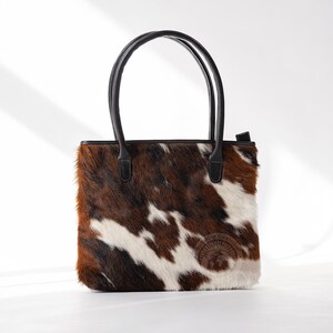 May include: A brown and white cowhide tote bag with black leather handles and a brown leather patch with the text "The Cowgirl" on the front.