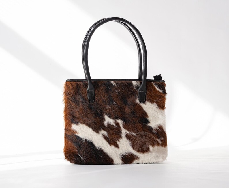 May include: A brown and white cowhide tote bag with black leather handles and a brown leather patch with the text "The Cowgirl" on the front.