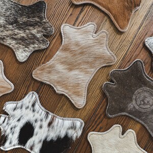 May include: A selection of cowhide coasters in shades of brown, tan, and black, displayed on a wooden surface. Each coaster features a distinctive pattern and shape, with a stitched edge. One coaster has a logo.