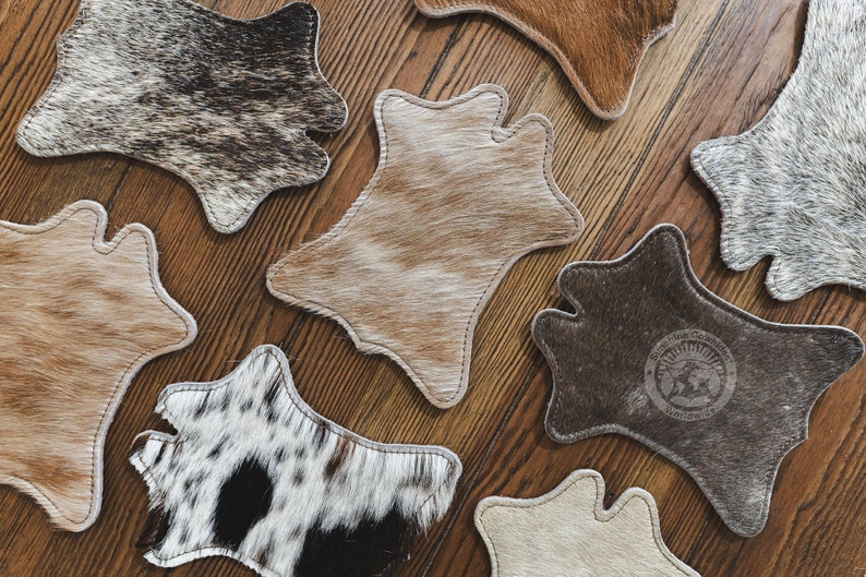 May include: A selection of cowhide coasters in shades of brown, tan, and black, displayed on a wooden surface. Each coaster features a distinctive pattern and shape, with a stitched edge. One coaster has a logo.