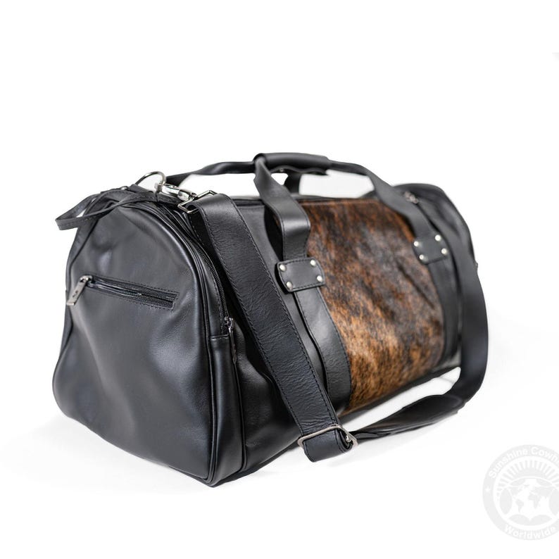 May include: A black duffel bag featuring smooth black leather-like side panels with a zippered pocket and a central section made of brown and black cowhide fur. The bag includes black leather handles, silver-toned metal rivets, and a shoulder strap.