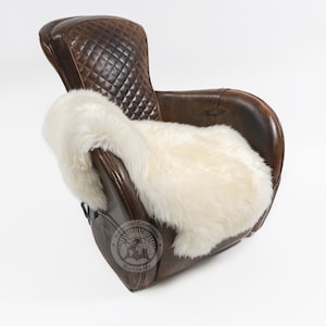 Puede incluir: Un sill&oacute;n de cuero marr&oacute;n con respaldo acolchado en forma de diamante y una manta de piel de oveja color crema sobre el asiento. La silla tiene una est&eacute;tica vintage y est&aacute; sobre un fondo blanco.