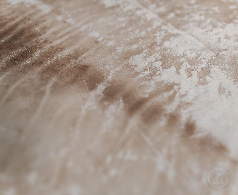 Puede incluir: Una vista de cerca de una superficie de tela con un patr&oacute;n texturizado y desgastado en tonos marr&oacute;n claro y blanco roto. El material muestra una apariencia suave, similar al terciopelo, con parches irregulares que crean un efecto envejecido.