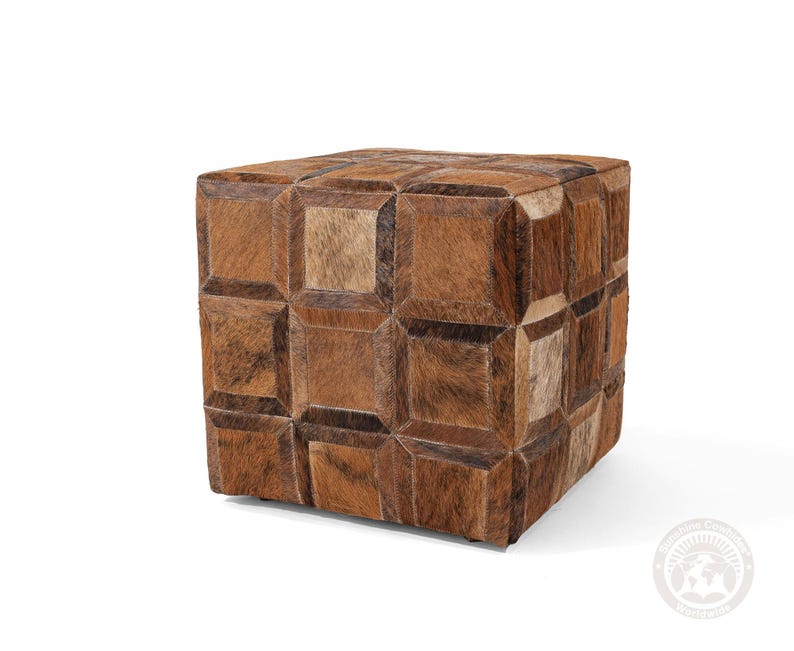 May include: A cube-shaped ottoman with a patchwork design. The ottoman is covered in brown and tan cowhide squares, creating a geometric pattern. The cowhide has a natural, textured appearance, adding visual interest to the piece. The ottoman is a decorative furniture item.