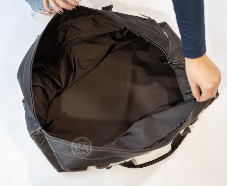 May include: A large dark brown leather travel bag with a spacious interior lined in dark brown fabric. The exterior features patches of white and brown cowhide fur. A circular logo with the text Sunshine Cowhides Worldwide is visible.