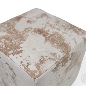 Puede incluir: Un puf de forma cuadrada con un acabado desgastado y un patr&oacute;n moteado en tonos blanco roto y marr&oacute;n claro. La superficie tiene una apariencia texturizada con costuras visibles a lo largo de los bordes, creando una forma de cubo con esquinas redondeadas.