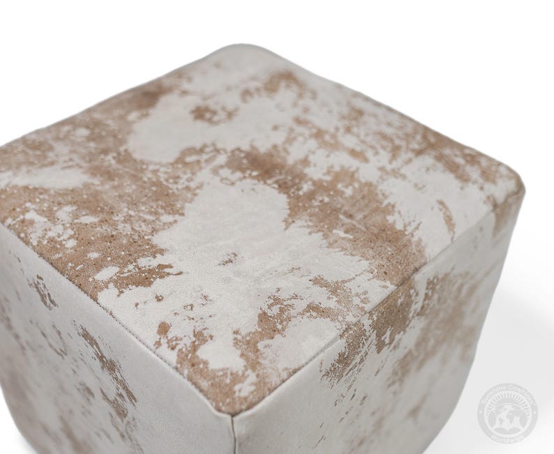 Puede incluir: Un puf de forma cuadrada con un acabado desgastado y un patr&oacute;n moteado en tonos blanco roto y marr&oacute;n claro. La superficie tiene una apariencia texturizada con costuras visibles a lo largo de los bordes, creando una forma de cubo con esquinas redondeadas.