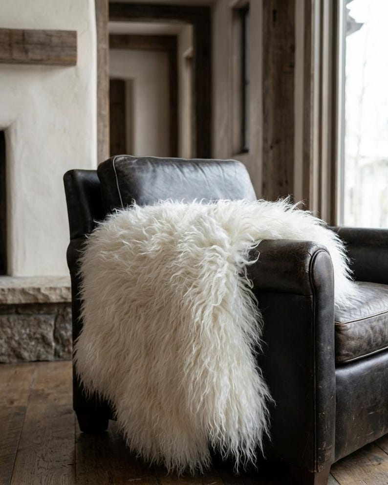 Puede incluir: Un sill&oacute;n de cuero marr&oacute;n oscuro con una manta de piel sint&eacute;tica blanca y esponjosa sobre &eacute;l. La manta tiene fibras largas y peludas. La silla est&aacute; en una habitaci&oacute;n con chimenea y detalles de madera.