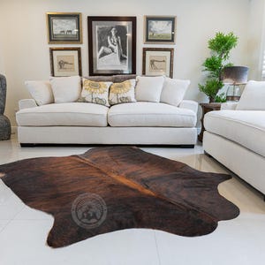 May include: A brown and white cowhide rug with a natural pattern. The rug is laid out on a white tiled floor in a living room setting with a white sofa and a white armchair.