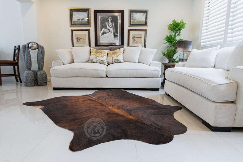 May include: A brown and white cowhide rug with a natural pattern. The rug is laid out on a white tiled floor in a living room setting with a white sofa and a white armchair.