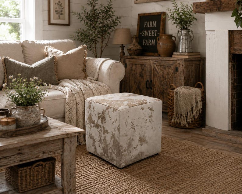 Puede incluir: Un taburete cuadrado con un patr&oacute;n desgastado y moteado en blanco y beige en su tapicer&iacute;a de tela. El mueble en forma de cubo se asienta sobre una alfombra tejida, mostrando una superficie texturizada con una est&eacute;tica neutra y envejecida.